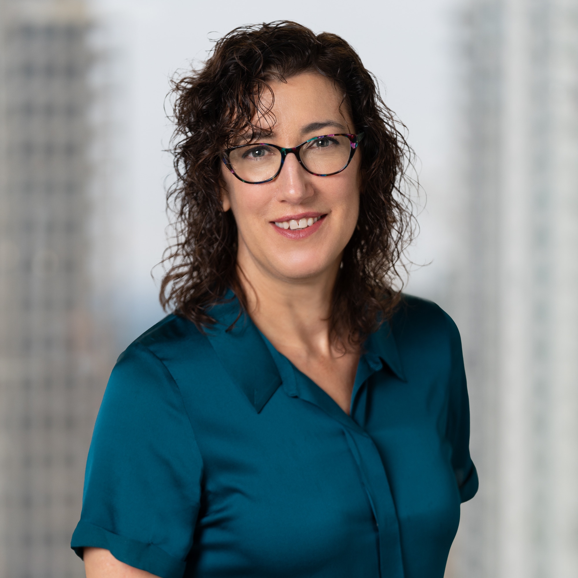 photo of Rebecca Molesworth - brown curly hair, black framed glasses, and a dark teal blouse against a blurred background of a window overlooking the city