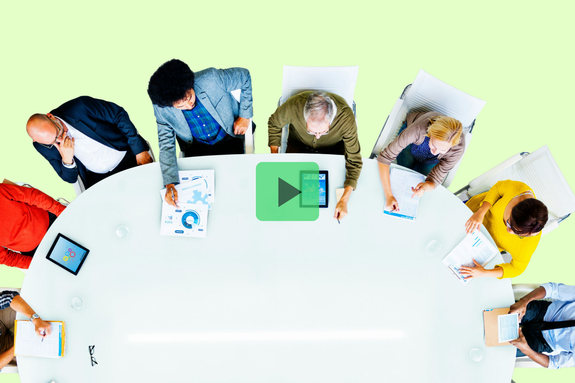 A group of people sit around an oval table with documents, laptops, and tablets, viewed from above, on a light green background. A green play button is overlaid on the image center.