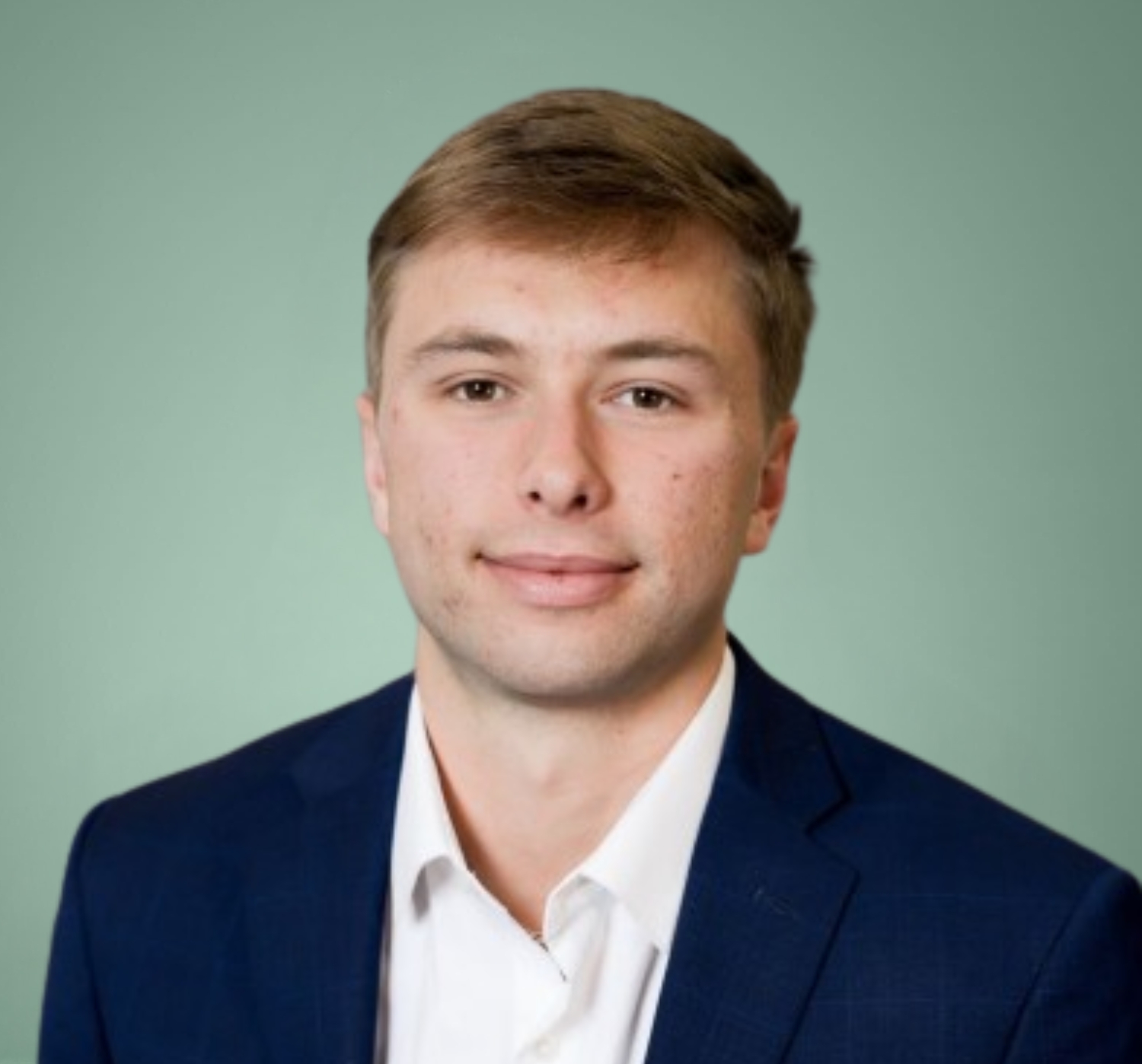 A young man with short light brown hair, wearing a white shirt and navy blazer, poses in front of a plain light green background.