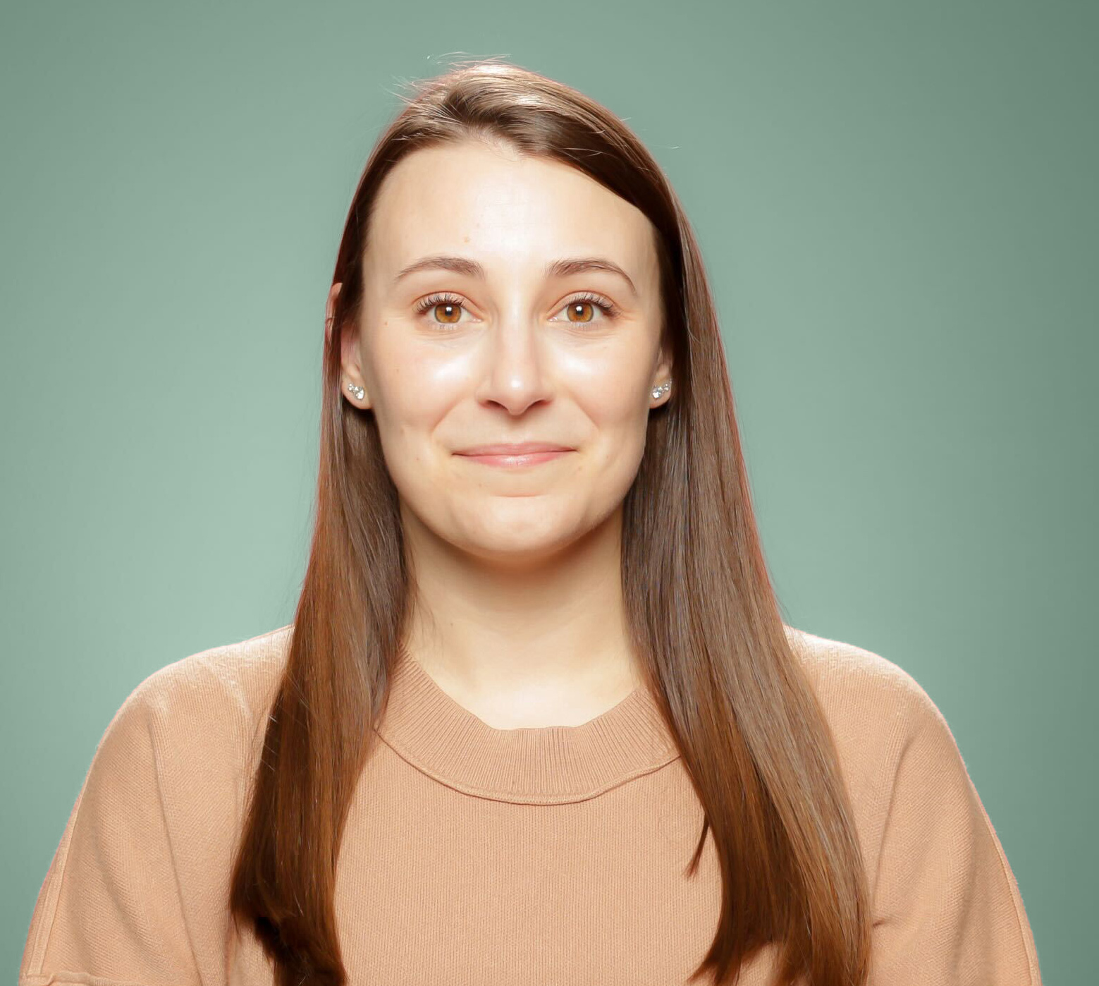 A woman with straight brown hair and a beige sweater poses in front of a plain green background, looking at the camera and smiling softly.
