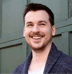A man with short dark hair and a trimmed beard smiles while standing in front of a green wooden wall, wearing a dark cardigan over a light shirt.
