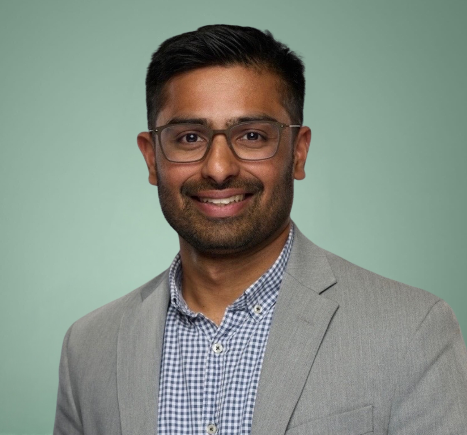 A man with short dark hair, glasses, and a trimmed beard wears a gray blazer over a checkered shirt, posing and smiling in front of a plain green background.