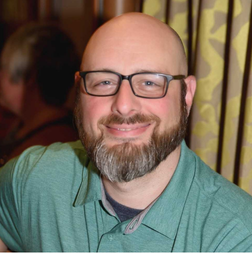 A smiling man with glasses, a bald head, and a trimmed beard, wearing a green collared shirt, sits indoors in front of patterned curtains.