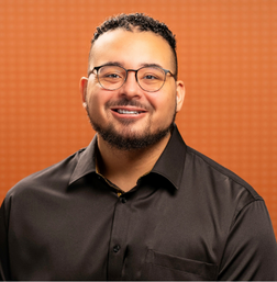 A man with short curly hair, glasses, and a trimmed beard smiles while wearing a dark button-up shirt, posed in front of an orange patterned background.