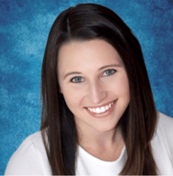 Woman with long brown hair and blue eyes smiling at the camera, wearing a white top, posed against a blue textured background.