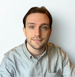 A young man with light skin and short brown hair, wearing a light gray button-up shirt, sits in front of a plain white background, facing the camera with a slight smile.