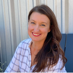 Woman with long brown hair and a plaid shirt smiles at the camera while sitting in front of a corrugated metal wall.