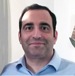 A man with dark hair, wearing a light blue collared shirt, stands indoors in front of a light-colored wall.