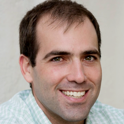 A man with short brown hair wearing a green and white checkered shirt smiles at the camera against a plain light background.