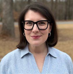A woman with short brown hair, wearing black glasses and a light blue collared shirt, stands outside with trees and a road in the background.