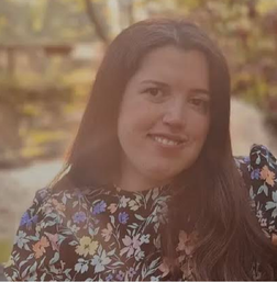 A woman with long brown hair and a floral top smiles outdoors with sunlight and trees in the background.