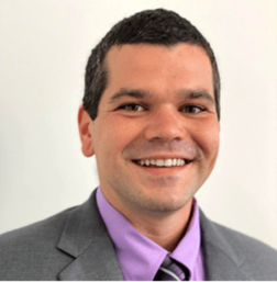 A man with short dark hair wearing a gray suit jacket, light purple dress shirt, and striped tie, smiling in front of a plain light background.