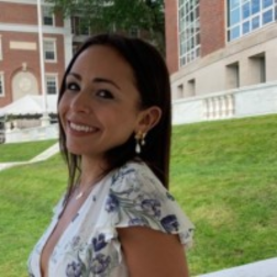 A woman with long dark hair in a floral dress smiles outdoors in front of a brick building and green lawn.