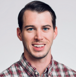 A young man with short brown hair and blue eyes, wearing a plaid shirt, smiling at the camera against a plain light background.