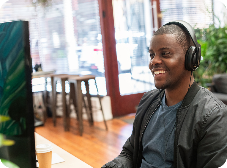 A man wearing headphones sits at a desk with a computer monitor, smiling. A coffee cup is on the desk and the background shows stools, plants, and large windows.