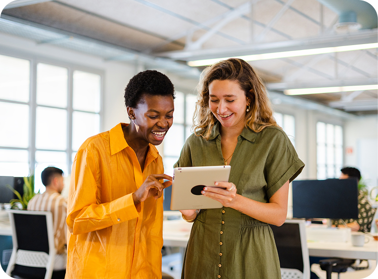 Two women standing in an office, smiling and looking at a tablet together. Other people are seated and working at desks in the background.