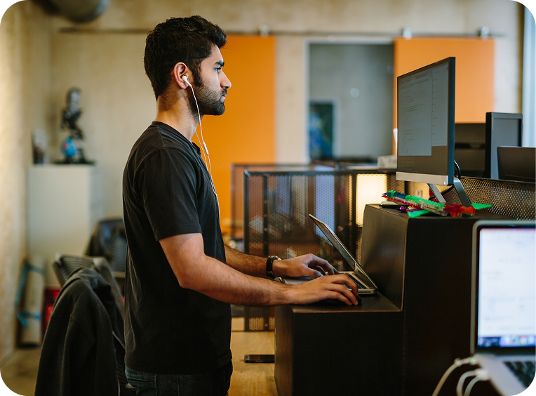 A man stands at a desk using a computer and a laptop, wearing earphones in an office setting.