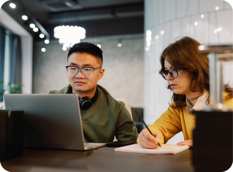 Two people sit at a desk; one works on a laptop while the other writes in a notebook. Both wear glasses and appear focused in a modern office setting.