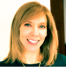 A woman with straight, shoulder-length light brown hair smiles at the camera. She is wearing a dark top and a gold necklace.