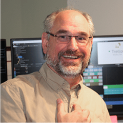 A man with glasses and a beard smiles at the camera, standing in front of computer monitors displaying various software and graphics.