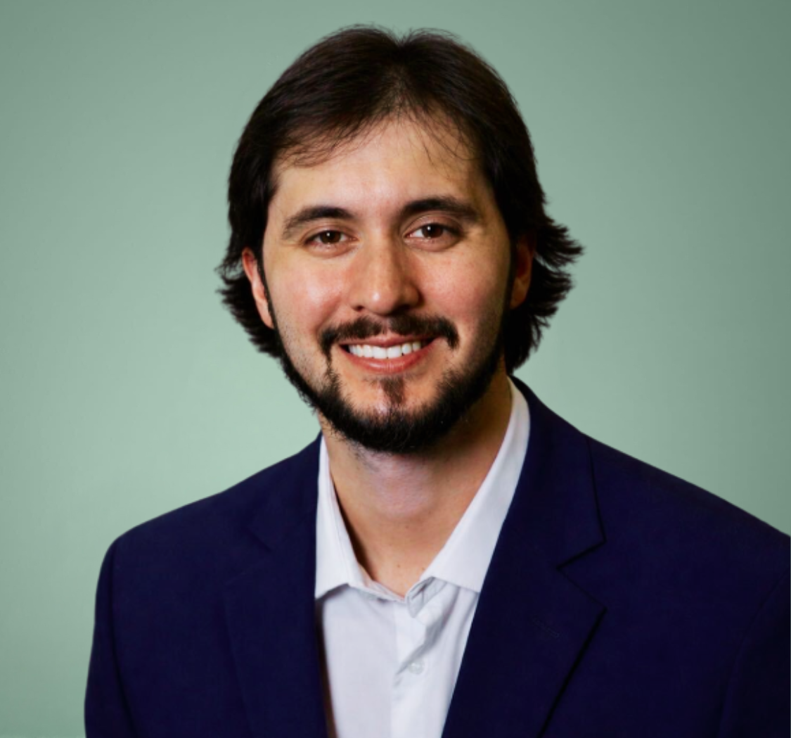 A man with dark hair and a beard, wearing a navy blazer and white shirt, smiles in front of a plain light green background.