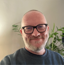 A man with a beard and glasses smiles at the camera in a well-lit room, with green plants visible in the background.