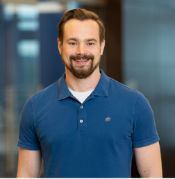 A man with short brown hair and a beard, wearing a blue polo shirt, stands indoors in front of a blurred office background.
