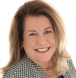 Smiling woman with shoulder-length light brown hair, wearing a black and white patterned jacket, posing in front of a plain white background.