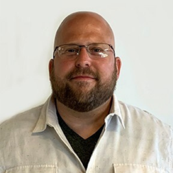 A man with a shaved head, beard, and glasses is wearing a white button-up shirt over a dark t-shirt, standing against a plain white background.