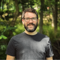 A man with glasses and a beard stands outdoors in front of green trees, wearing a dark gray t-shirt and smiling at the camera.