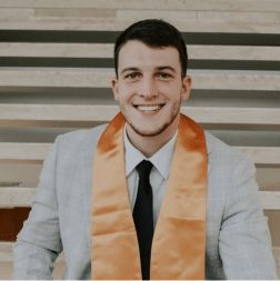 A young man in a light gray suit and black tie wears an orange graduation stole, smiling while seated in front of light-colored steps.