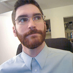 A man with glasses and a trimmed beard sits indoors, wearing a light blue collared shirt, looking slightly upwards to his right. Bookshelves are visible in the background.