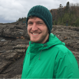 A smiling person in a green jacket and dark knit hat stands outdoors on rocky terrain with trees in the background.