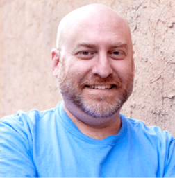 A bald man with a beard and mustache smiles while standing against a textured light brown wall, wearing a light blue t-shirt.
