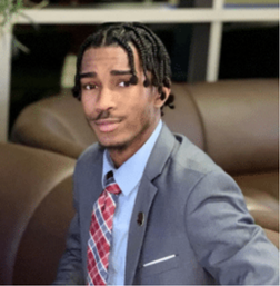 A young man in a gray suit and plaid tie sits on a brown couch, looking at the camera with a neutral expression.