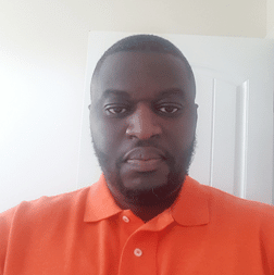 A man with a short beard wearing an orange polo shirt stands indoors in front of a white door, looking directly at the camera.