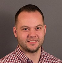 A man with short brown hair and a trimmed beard wearing a red and white checkered shirt, smiling slightly in front of a plain gray background.