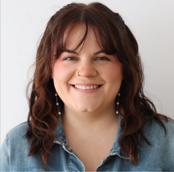 A woman with wavy brown hair and bangs smiles at the camera. She is wearing a denim shirt and dangly earrings, standing against a plain light background.