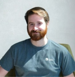 A man with a beard and mustache sits on a green chair, wearing a gray t-shirt with a small logo, against a plain light background.