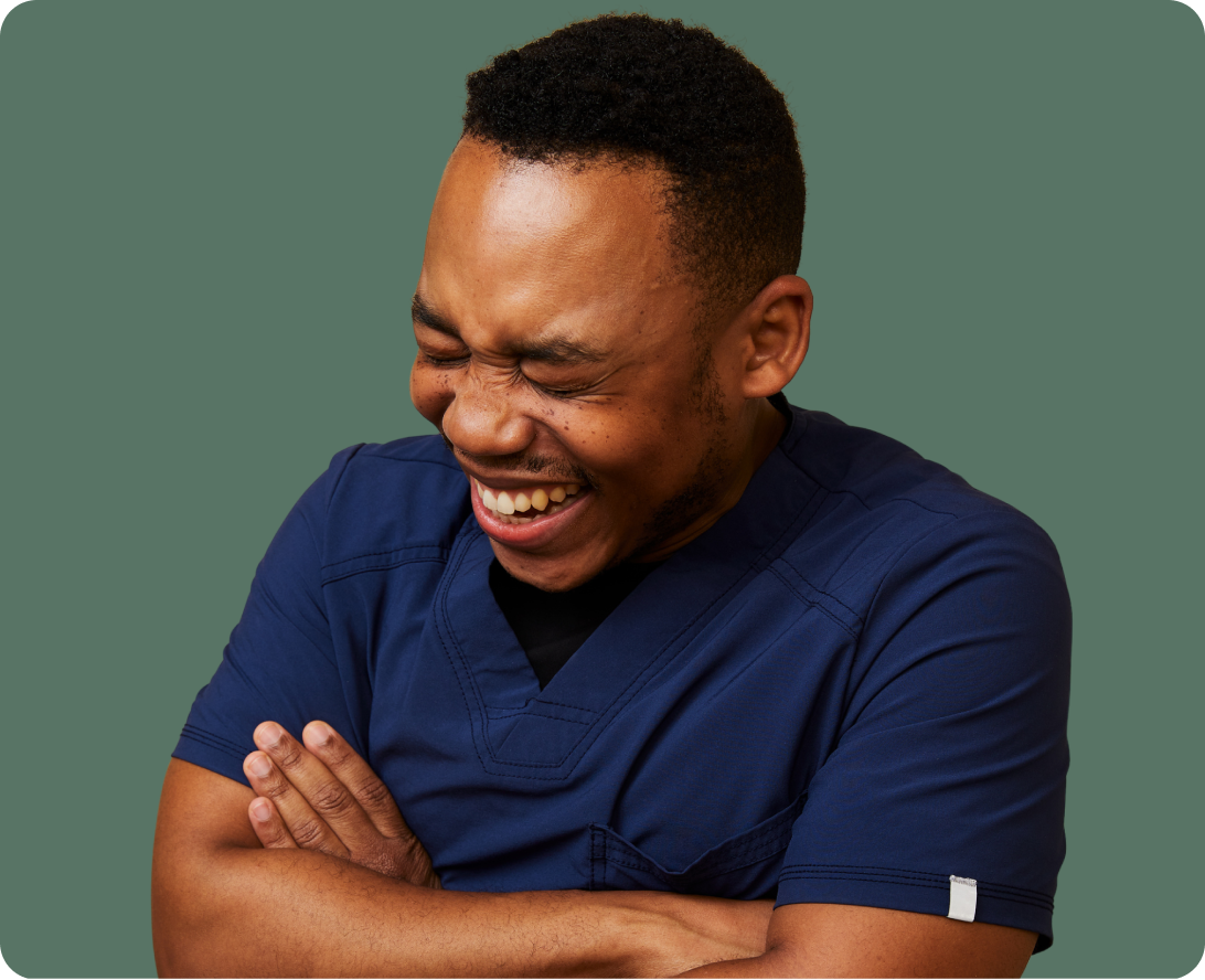 A person wearing navy blue scrubs stands with arms crossed, eyes closed, and laughing against a plain green background.