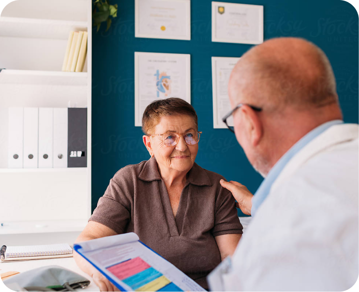 Older woman sits across from a doctor in an office, listening as he reviews a clipboard with documents, with certificates visible on the wall behind them.