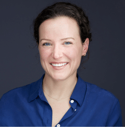 A woman with curly brown hair, wearing a blue collared shirt, smiles at the camera against a dark gray background.