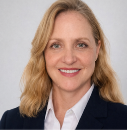 A woman with blonde hair wearing a navy blazer and white shirt, smiling at the camera against a plain light background.