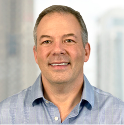 A man with short gray hair and a light blue collared shirt smiles at the camera. A cityscape is blurred in the background.
