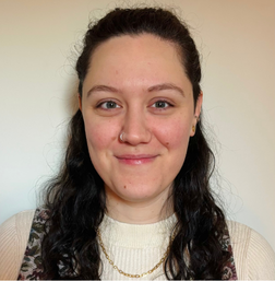 A woman with long dark curly hair, wearing a cream top, a floral-patterned vest, and a gold necklace, smiles at the camera against a plain background.