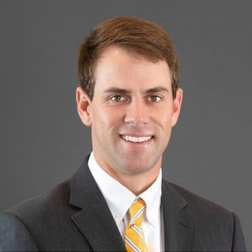A man in a dark suit, white shirt, and yellow striped tie poses for a professional headshot against a plain gray background.