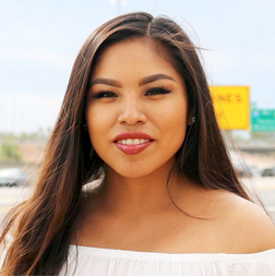 A young woman with long brown hair and a white off-the-shoulder top stands outdoors, smiling at the camera. Road signs and a blurred highway are visible in the background.