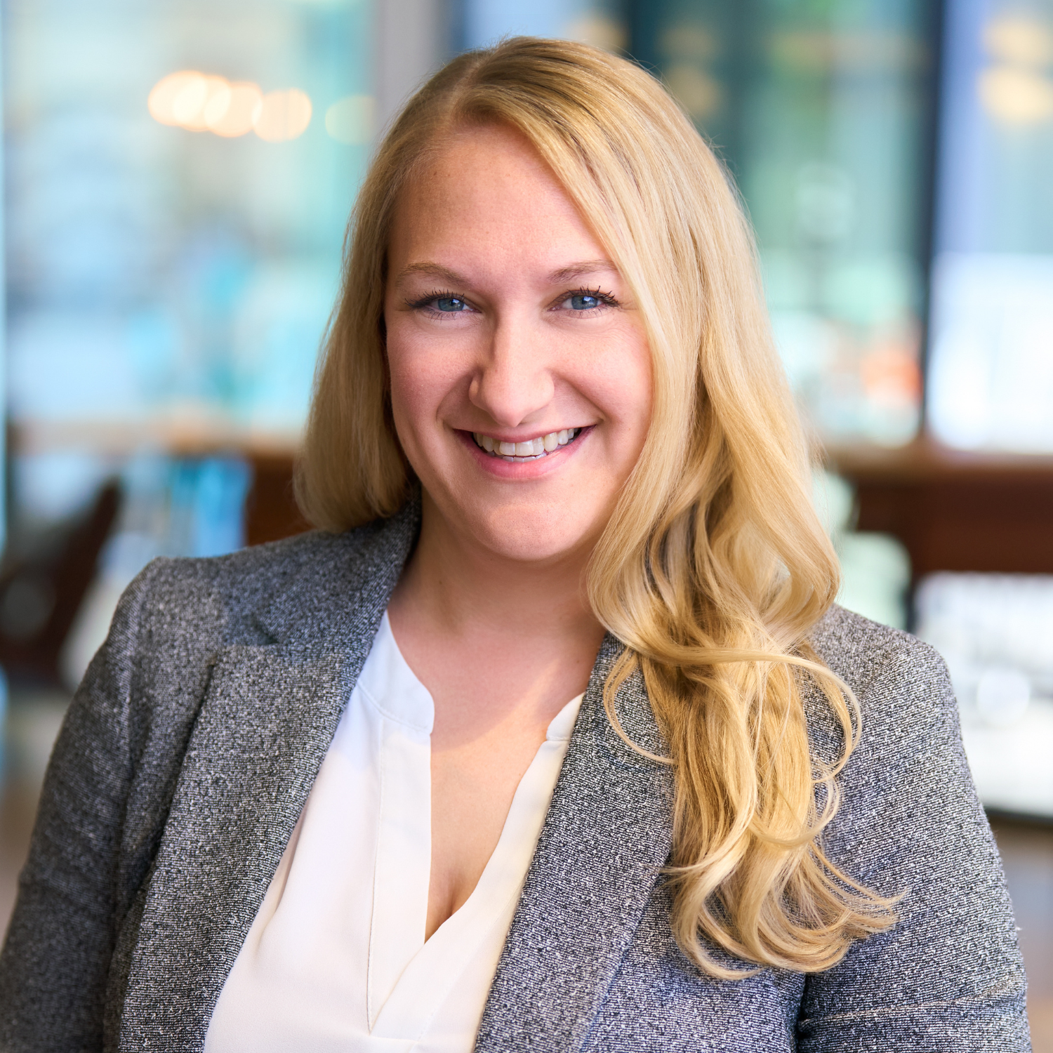 A woman with long blonde hair wearing a gray blazer and white blouse smiles at the camera in a bright office setting.