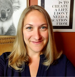 Woman with blonde hair smiling at the camera, sitting indoors with a framed koala picture and a motivational quote on the wall behind her.