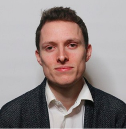 A man with short brown hair wearing a white shirt and dark blazer poses in front of a plain light background.